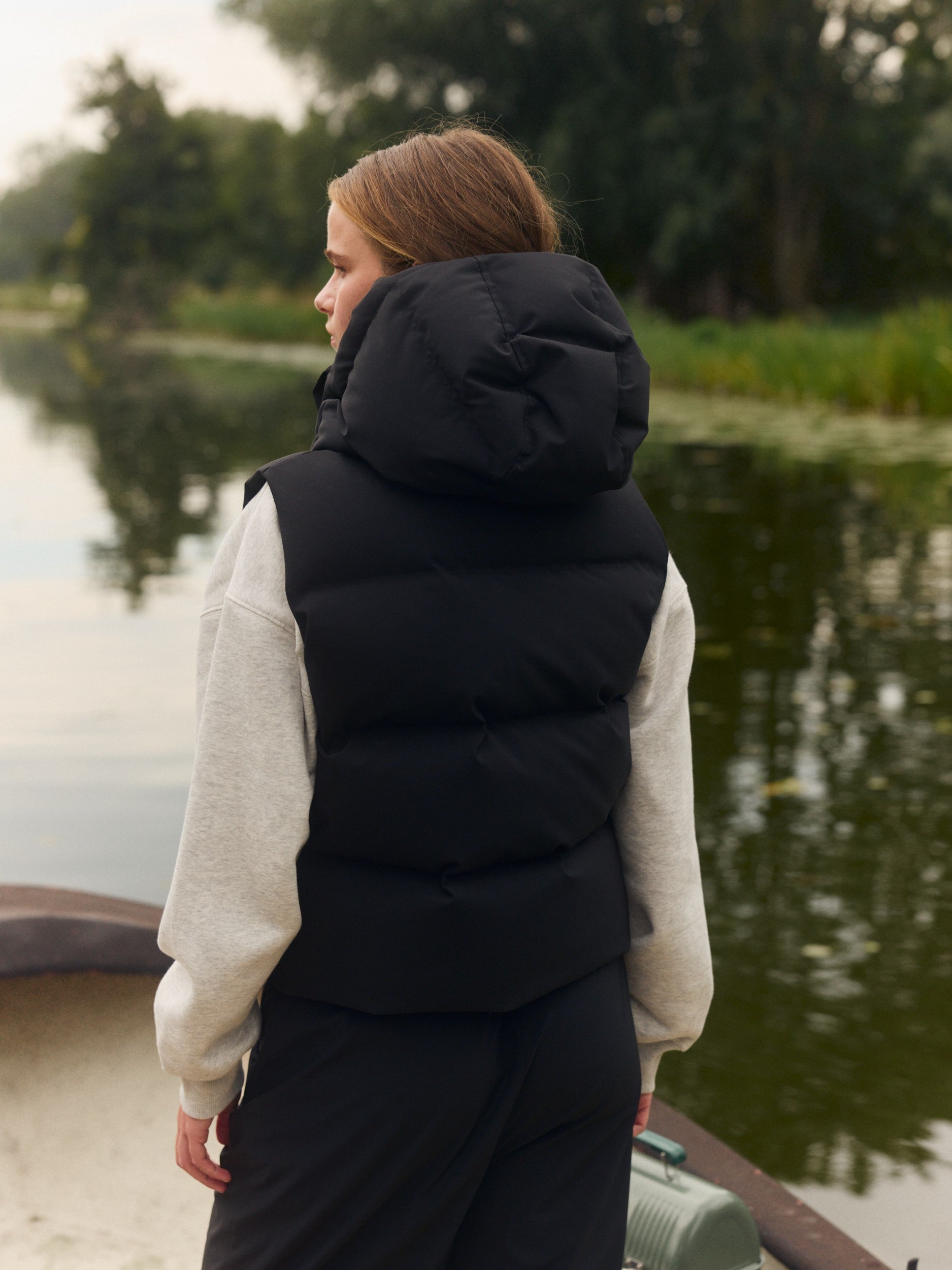 Woman wearing a black hooded puffer vest over a grey sweatshirt outdoors by a lake.